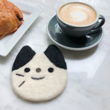 A needle felted tuxedo cat coaster, part of a set, rests on a white marble table beside a coffee cup and pastry, creating a relaxed scene. This round, 4-inch handcrafted wool felt coaster features a white cat face with black markings on its ears and head. Simple stitched black eyes, a brown nose, a curved black mouth, and whiskers define its charming expression. Ethically handmade from cozy sheep wool, it offers durable heat resistance for surfaces.