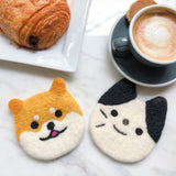 Two handcrafted Felt Tuxedo Cat Coasters rest on a marble table beside a chocolate pastry and a cup of coffee, creating a cozy and inviting scene. Each wool felt coaster is round with small, perky ears and features stitched eyes, a nose, and a mouth, portraying a cheerful expression. One distinct coaster is black and white, highlighting its unique design.