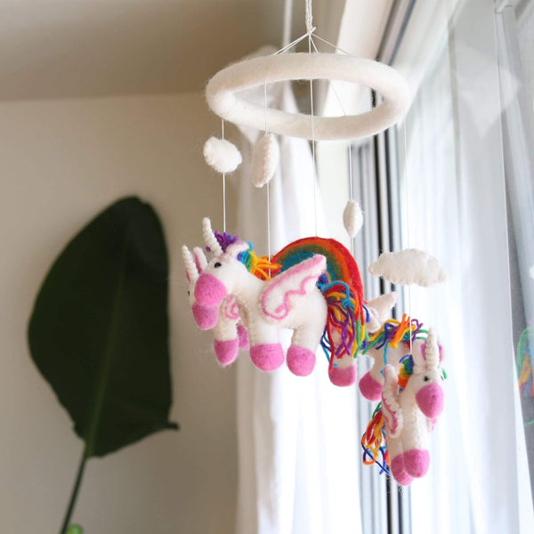 A felt unicorn baby mobile hangs in front of a window, suspended by strings from a circular ring. The mobile features two unicorn figures with rainbow manes and tails, one appearing larger due to perspective. Made from 100% New Zealand wool felt, each figure is needle felted individually for a 3D design.