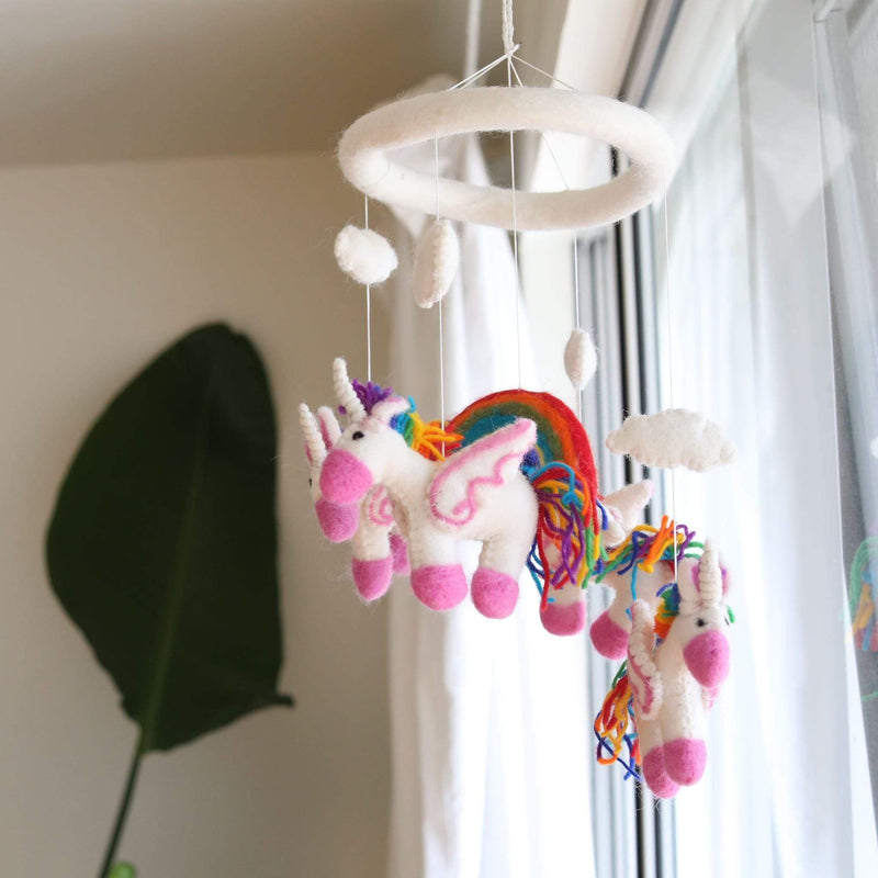 A felt unicorn baby mobile hangs in front of a window, suspended by strings from a circular ring. The mobile features two unicorn figures with rainbow manes and tails, one appearing larger due to perspective. Made from 100% New Zealand wool felt, each figure is needle felted individually for a 3D design.