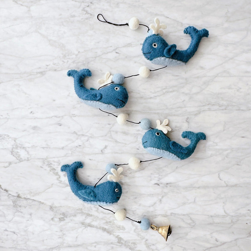 A charming wool felt whale garland rests on a white marble surface, creating a cheerful atmosphere. This felt garland features four blue, needle-felted whales, each with a cheerful smile, a white water spout, and a light gray belly. They are connected by a black string, interspersed with white and light blue felt balls, with a small copper bell dangling playfully at the bottom.