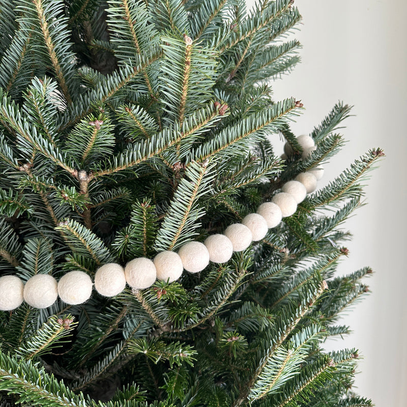 A Felt White Pompom Ball Garland made of wool felt is draped horizontally across the branches of a lush, green Christmas tree, evoking a cozy winter holiday atmosphere. The garland features evenly spaced, round white felt balls with a soft texture, nestled among the dark green evergreen needles, providing a gentle contrast.