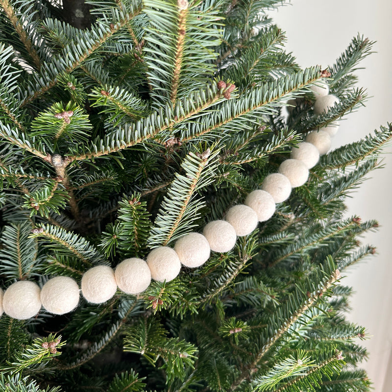 A White Pompom Ball felt garland is draped across the green branches of a Christmas tree in a close-up shot. The festive felt garland features a series of evenly spaced, round, off-white felt pompom balls, creating a cozy winter holiday ambiance.