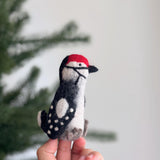 A vibrant, handcrafted felt woodpecker finger puppet is held by a hand against a blurred background featuring a Christmas tree. This unique wool felt finger puppet showcases a striking red head, a black and white body, a black beak, and a distinct crest. Its detailed textures highlight the exquisite, handmade quality, adding playful charm to any setting.