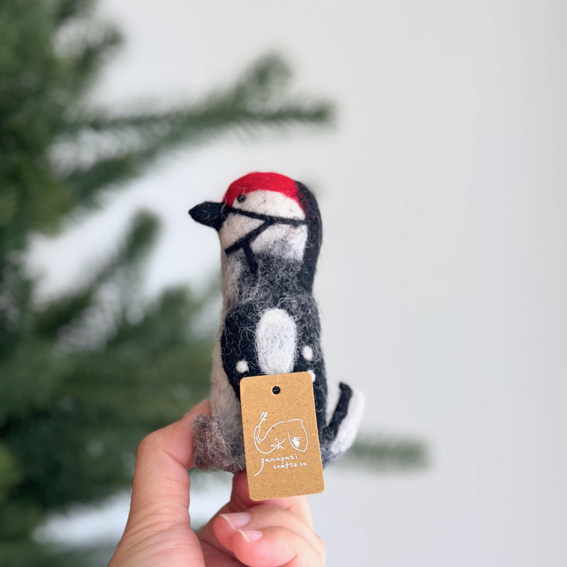 A wool felt woodpecker finger puppet is gently held by a female hand, presented against a soft, natural backdrop with a blurred green Christmas pine tree. This handcrafted wool felt kids toy showcases detailed black, white, and red felt work, capturing the bird's distinctive crest and soft texture. The scene exudes an artisanal and seasonal charm.