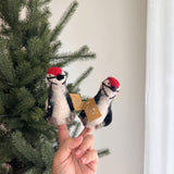 A handcrafted, needle-felted wool felt woodpecker finger puppet is shown worn on a hand, set against a blurred green Christmas tree. This unique felt toy for kids features a black and white body with vibrant red accents and a distinctive crest. Its eco-friendly design embodies whimsical craftsmanship.
