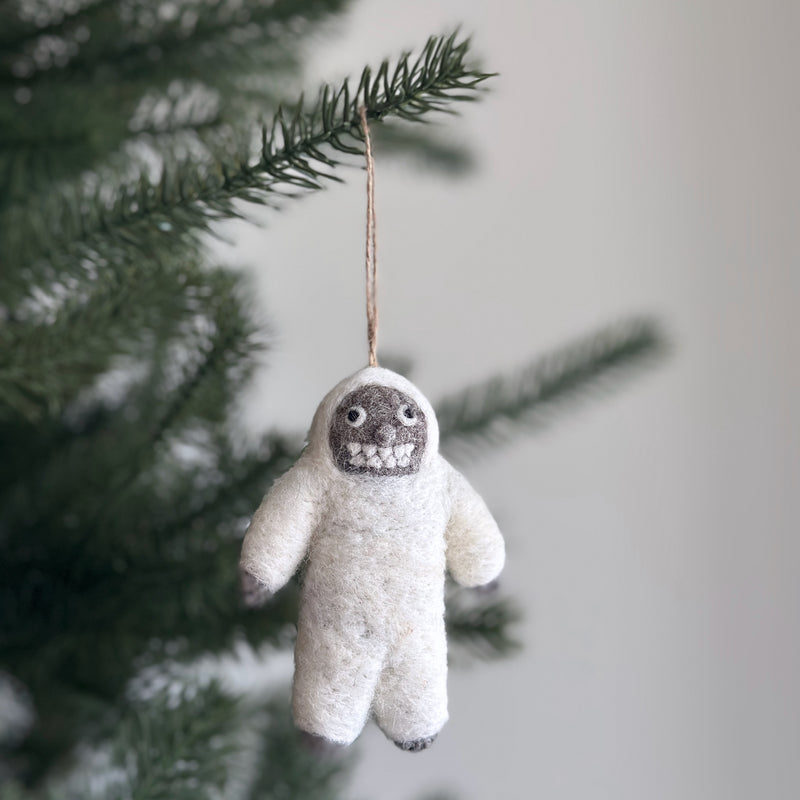 A white wool felt yeti ornament hangs from a branch of a lush green Christmas tree, creating a joyful and festive atmosphere. This needle-felted yeti ornament features a fluffy white body and a contrasting dark grey face with expressive detailing. A jute string suspends the whimsical and cozy yeti Christmas ornament.