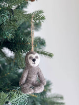 A gray felt yoga sloth ornament, suspended by a jute string, hangs from a Christmas tree branch. The unique Christmas ornament features a white face, a small black nose, and a small black mouth. It is depicted in a meditative seated pose with its eyes gently closed, creating a peaceful and festive mood.