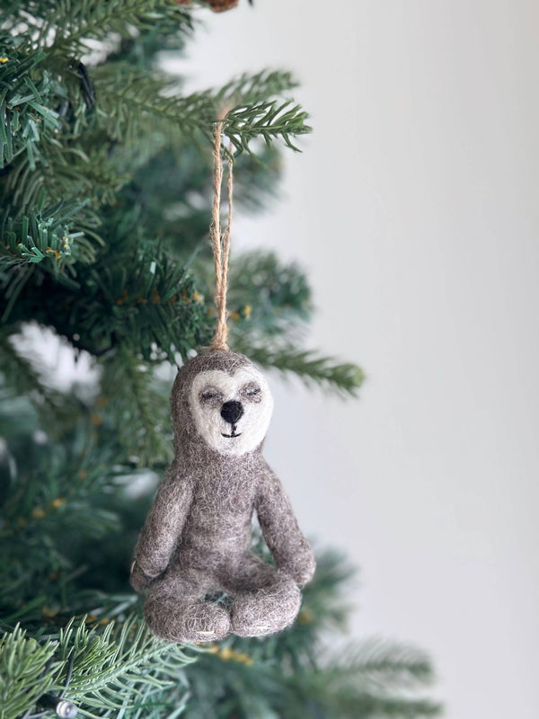 A gray felt yoga sloth ornament, suspended by a jute string, hangs from a Christmas tree branch. The unique Christmas ornament features a white face, a small black nose, and a small black mouth. It is depicted in a meditative seated pose with its eyes gently closed, creating a peaceful and festive mood.