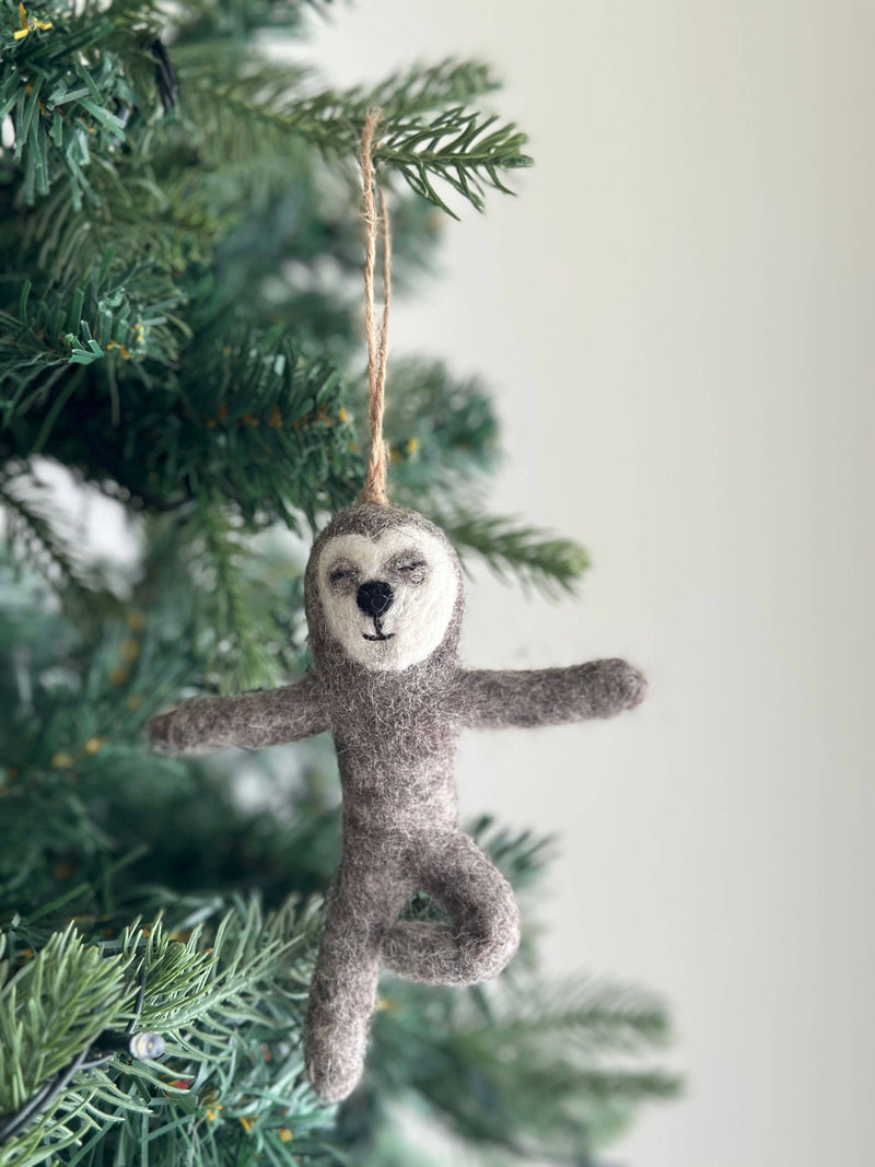 A handmade felt Yoga Sloth ornament hangs serenely from a lush green Christmas tree branch. This charming gray felt sloth, with a white face, black nose, and closed eyes, features flexible wire limbs posed whimsically with outstretched arms and legs. A jute string is attached to its head, allowing it to gracefully adorn the tree, adding a peaceful and festive touch.