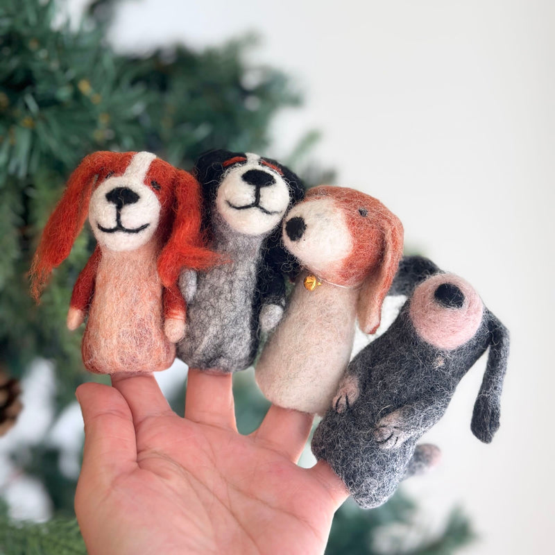A female hand holds four unique, handcrafted felt dog finger puppets on its fingertips, presented against a blurred Christmas tree background. These handmade felt dog finger puppets come in assorted colors: reddish-brown and cream, grey and black, cream and reddish-brown with a gold bell, and grey with pink accents. Each features a smiling face with stitched details, highlighting their playful, one-of-a-kind design.