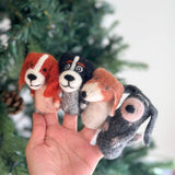 Four assorted wool felt dog finger puppets are held by a female hand against a blurred festive Christmas tree background. These unique handcrafted felt kids toys, made by female artisans, feature diverse designs: one with long orange ears and a white face stripe, another with black ears and a white face, a third with short brown ears and a white stripe, and a gray one with a single pink ear.
