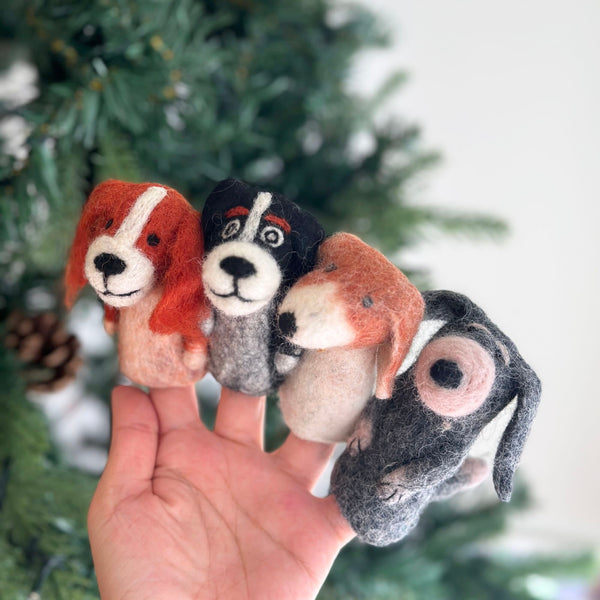 Four assorted wool felt dog finger puppets are held by a female hand against a blurred festive Christmas tree background. These unique handcrafted felt kids toys, made by female artisans, feature diverse designs: one with long orange ears and a white face stripe, another with black ears and a white face, a third with short brown ears and a white stripe, and a gray one with a single pink ear.