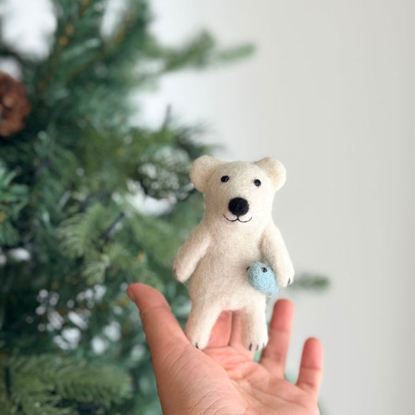 A handmade needle felted polar bear finger puppet, crafted from white wool felt and depicting a polar bear catching a small blue fish, is held in a female hand. The charming polar bear has small black eyes, a black nose, and a black mouth, displaying a cheerful expression. The blurred Christmas tree backdrop creates a festive and playful atmosphere for this unique finger puppet.