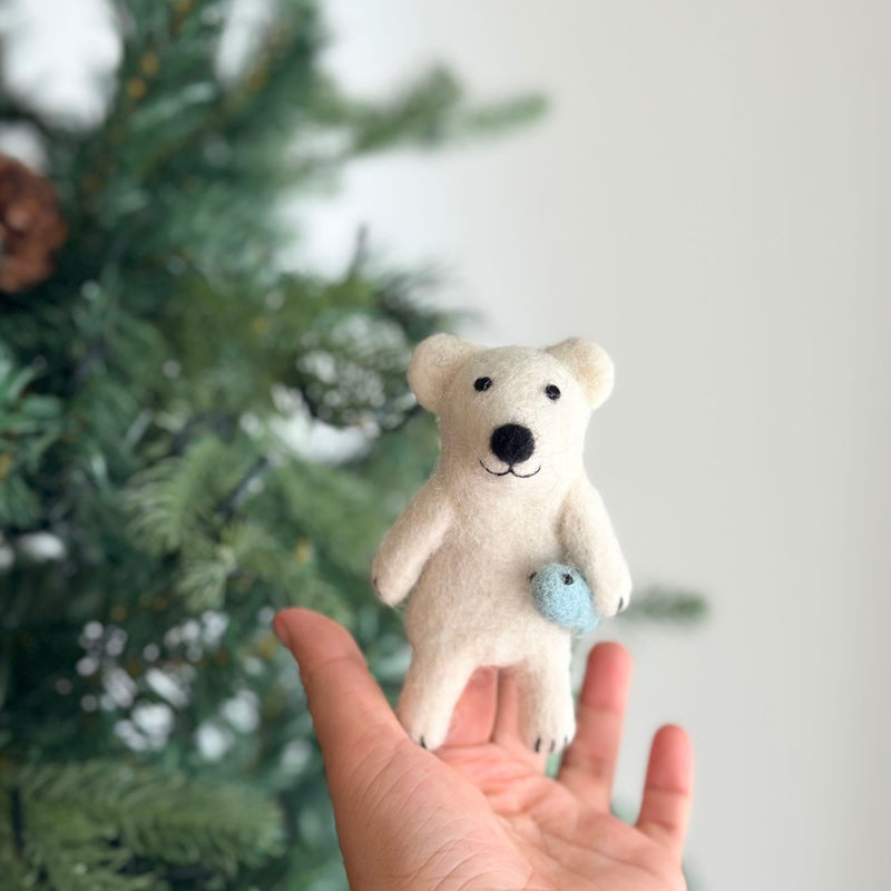 A handmade needle felted polar bear finger puppet, crafted from white wool felt and depicting a polar bear catching a small blue fish, is held in a female hand. The charming polar bear has small black eyes, a black nose, and a black mouth, displaying a cheerful expression. The blurred Christmas tree backdrop creates a festive and playful atmosphere for this unique finger puppet.