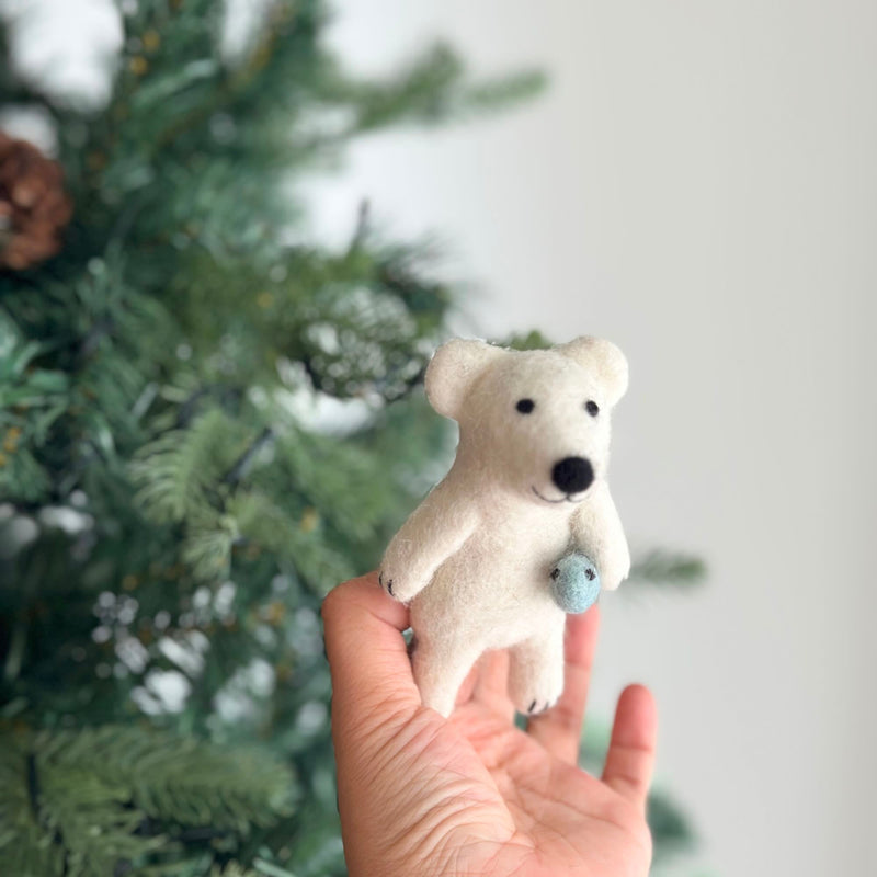 A handmade felt polar bear finger puppet, shown catching a fish, is held in a female hand against a green Christmas tree with a pine cone. The white polar bear features black eyes and nose, rounded ears, and visible paws, holding a round light blue fish in one paw. This unique felt toy for kids evokes imaginative play.