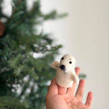 A handmade felt seal finger puppet is held in an open female hand, set against a blurred green background resembling a Christmas tree. This charming white seal features a rounded body with flippers, black eyes, a nose, and whiskers, ideal for imaginative storytelling.
