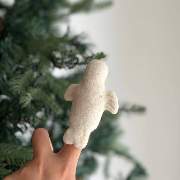 A playful, off-white wool felt seal finger puppet is held in a female hand, set against a soft-focused festive backdrop of green pine needles resembling a Christmas tree. This handcrafted felt seal features a rounded body and flippers, embodying a charming, simple design.