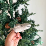 A light beige wool felt shark finger puppet is held by a female hand. This handmade puppet features a tail, fins, and stitched details for a friendly mouth and eyes, perfect for imaginative play.