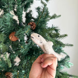 A handmade felt shark finger puppet is held upright by a hand, set against a blurred Christmas pine tree background adorned with pine cones. This light gray shark finger puppet features a streamlined body, tiny black eyes, and stitched gills. Its simple, soft texture highlights its unique, handcrafted design.