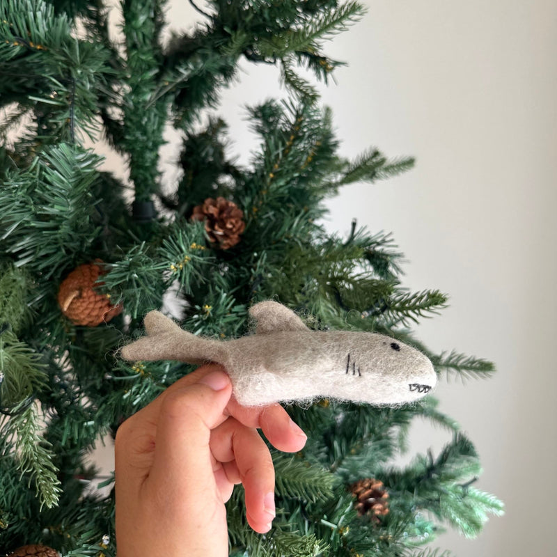 A cheerful felt shark finger puppet, handcrafted from light gray felt, is gently held by a hand against a festive Christmas tree adorned with pine cones. This handmade finger puppet features a rounded body, a dorsal fin, side fins, a tail, black eyes, and a stitched mouth with visible teeth, perfect for holiday play.
