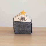 A handcrafted wool felt stable, part of a Nativity Scene Finger Puppet Story Set, rests on a simple wood-grain surface against a white backdrop. This unique gray stable features a cream-colored roof adorned with brown thread accents and a yellow star, plus a stitched gray window on the front. Its visible white stitching highlights the handmade felt construction, ideal for storing your felt toys for kids.