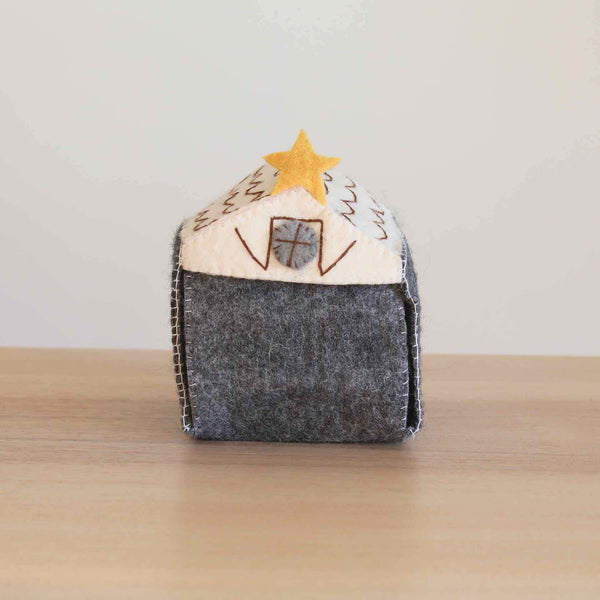 A handcrafted wool felt stable, part of a Nativity Scene Finger Puppet Story Set, rests on a simple wood-grain surface against a white backdrop. This unique gray stable features a cream-colored roof adorned with brown thread accents and a yellow star, plus a stitched gray window on the front. Its visible white stitching highlights the handmade felt construction, ideal for storing your felt toys for kids.