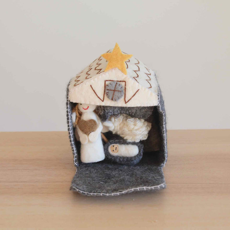 A handmade felt finger puppet set, featuring a nativity scene with 12 intricately crafted figures, including Baby Jesus, Mary, Joseph, three kings, an angel, a shepherd, a cow, a sheep, and a donkey. The set is presented in a felt stable with an open door, placed on a wooden table with a white surface, evoking comfort and tradition.