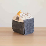 A charming, handcrafted felt nativity scene stable, part of a Finger Puppet Story Set—Nativity Scene, rests on a light wood surface. The dark gray stable features a white roof with brown scalloped tiles and a yellow star above a square window. Visible white stitching highlights the handmade quality of this unique felt piece, designed for storing nativity scene finger puppets.