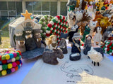 A handmade nativity scene felt finger puppet set is displayed on a table, creating a traditional holiday atmosphere. The 12 intricately detailed felt finger puppets include baby Jesus in a manger, Mary, Joseph, three kings, an angel, a donkey, a sheep, and additional animals, all arranged around a stable background.