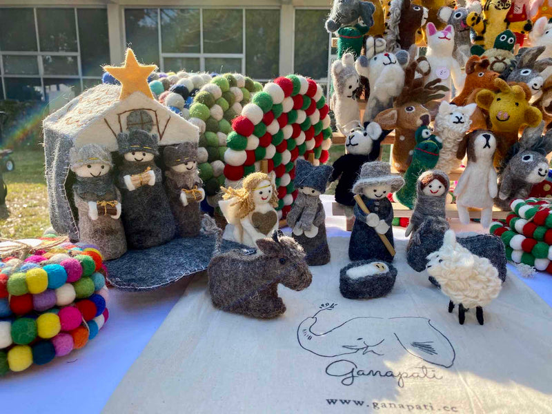 A handmade nativity scene felt finger puppet set is displayed on a table, creating a traditional holiday atmosphere. The 12 intricately detailed felt finger puppets include baby Jesus in a manger, Mary, Joseph, three kings, an angel, a donkey, a sheep, and additional animals, all arranged around a stable background.
