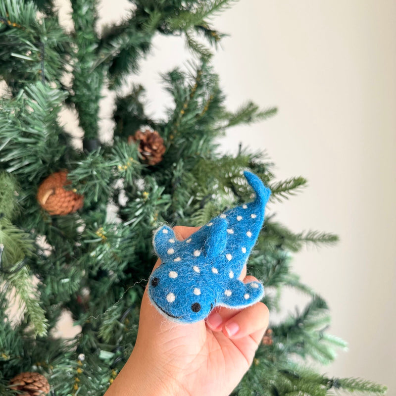 A handcrafted felt whale shark finger puppet is held in a female hand, displayed against a festive Christmas tree adorned with pine cones. This unique Starry Whale Shark puppet features white spots resembling a starry night sky, a small fin, and a tail. This handmade felt toy encourages imaginative play and oceanic adventures.