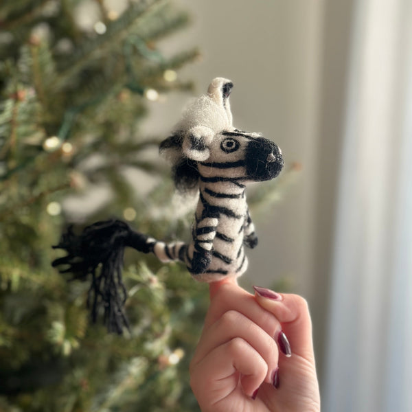 A handcrafted needle-felted zebra finger puppet is playfully presented, held by a female hand with dark red nails against the soft, festive glow of a Christmas tree. This unique felt puppet features iconic black and white stripes, a tufted mane, and a black tail, embodying a cozy holiday spirit.