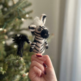 A handcrafted needle-felted zebra finger puppet, with detailed black and white stripes, a fluffy white mane, and a black tail, is displayed near a softly lit Christmas tree. This unique felt toy for kids evokes a playful and whimsical mood.
