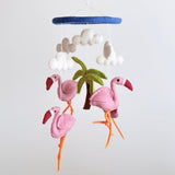 A handmade wool felt Flamingo Baby Mobile hangs from a white string against a plain background, featuring three flamingo figures with distinct poses and color variations in shades of pink and orange. The mobile includes a small felt palm tree between the flamingos, conveying a tropical theme and soft texture.