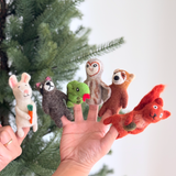 A female hand displays a unique set of six handcrafted forest animal wool felt finger puppets against a blurred Christmas tree backdrop. This charming collection includes a white bunny holding an orange carrot, a brown raccoon, a green snake with a red tongue, a light brown owl, a red panda, and a reddish-brown squirrel with a brown nut. Each felt finger puppet showcases intricate details.