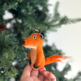 A handcrafted wool felt fox finger puppet is held upright by a female hand, set against a blurred Christmas pine tree background. This meticulously crafted, orange fox features a long, bushy tail with a white tip, small blue eyes, a black nose, and black ears with white tips. Its cream-colored belly and front paws, appearing held together, enhance its unique charm. This fair-trade, handmade toy supports female artisans.