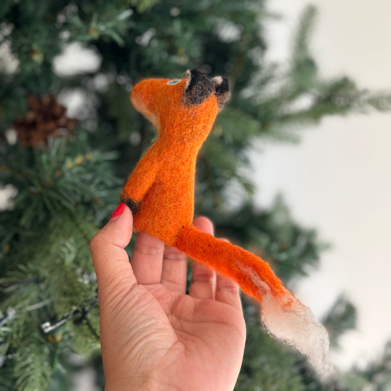 A female hand holds a vibrant orange handmade wool felt fox finger puppet, playfully set against a blurred Christmas pine tree backdrop. This charming felt fox puppet features a long, textured tail with a white tip, and dark accents on its ears and paws, evoking a cheerful mood.