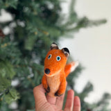 A wool felt fox finger puppet is playfully held by a hand, set against a blurred, festive Christmas tree background. This handcrafted felt toy features a vibrant orange body with a light-colored belly and a white-tipped tail. It has round blue eyes and black accents on its nose, ears, and paws, showcasing its slightly textured wool felt surface.