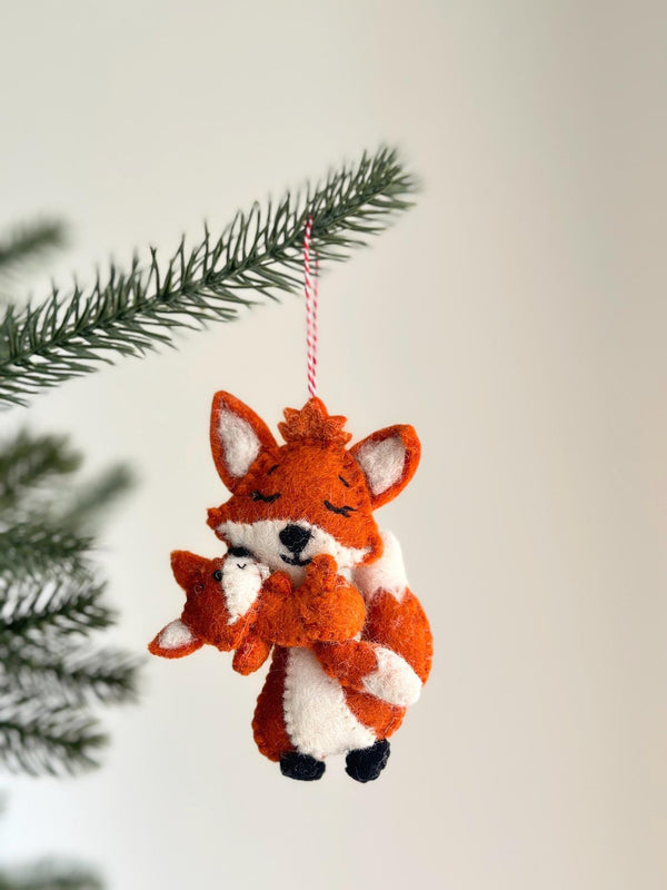 A handcrafted wool felt fox holding baby ornament is displayed on a pine branch against a bright white background. The orange mother fox, with a white chest and ear insides, lovingly embraces her smaller baby fox. Both felt foxes feature stitched closed eyes and petite black noses, with a red and white jute thread attached for hanging this charming Christmas ornament.