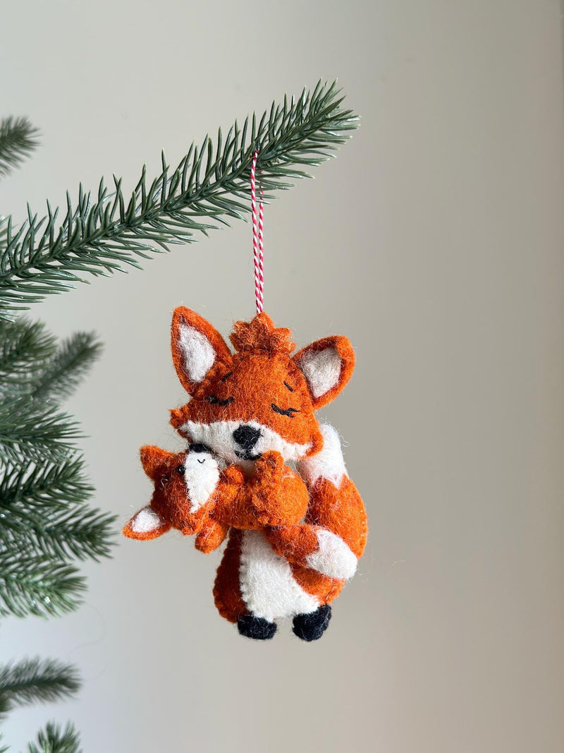 A heartwarming needle felted fox holding baby ornament hangs from a lush green Christmas tree branch, set against a neutral background. Crafted from soft orange and white wool felt, this unique felt ornament depicts a larger fox gently embracing a smaller one. Both foxes feature tiny perky ears, closed eyes, and subtle black stitched details, with a red and white jute thread attached for hanging.