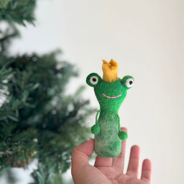 A handmade felt frog finger puppet is held in a female hand, set against a blurred Christmas tree, creating a festive scene. This 3D-designed, needle-felted wool felt frog finger puppet is green with a smiling pink mouth, white eyes, and a golden crown. Two small green felted balls hang from its sides, highlighting its charming details. This unique finger puppet is perfect for imaginative play or storytelling.