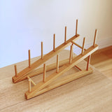 A light bamboo finger puppet shelf, part of a Wool Felt Finger Puppet Set, is cleanly displayed against a soft white background. The two-level, handcrafted bamboo shelf features dowels designed to neatly organize and showcase the included wool felt finger puppets. This fair-trade and environmentally friendly set offers a functional and aesthetically pleasing storage solution for kids' felt toys.