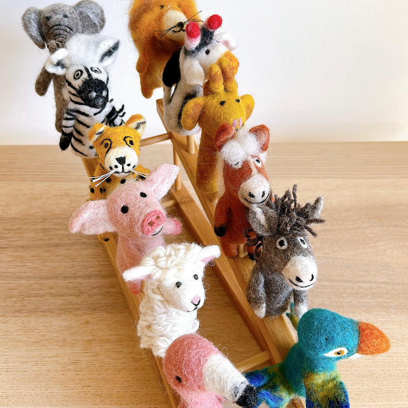 A colorful Wool Felt Finger Puppet Set, featuring 12 unique handcrafted animal puppets, is arranged on a light bamboo shelf against a plain white backdrop and light wood surface. The set includes needle felted elephant, zebra, tiger, pig, sheep, flamingo, parrot, donkey, horse, giraffe, mouse, and lion puppets. Each handmade wool felt animal displays distinct colors, shapes, and textures, creating a vibrant and cheerful collection.