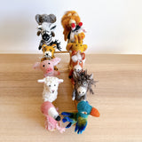 A collection of handmade wool felt finger puppets displayed on a bamboo shelf against a white background, featuring a giraffe, elephant, zebra, fox, sheep, cow, pig, and dog. Each puppet is unique in color and pattern, with some having additional details like ears or tails. The overall mood is playful and whimsical, suggesting fun and creativity associated with these finger puppets.