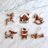 Six unique needle felted gingerbread ornaments, mimicking classic gingerbread cookies, are artfully arranged on a bright marble surface. This charming set includes a reindeer with white snowflakes, a gingerbread house with a bird, a rocking horse, a dachshund, a bear with a red bow, and a shark. Each handcrafted felt ornament is brown with intricate white needle-felted details and hangs from a jute string, adding a cozy, holiday feel.