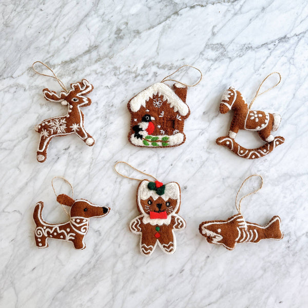 Six unique needle felted gingerbread ornaments, mimicking classic gingerbread cookies, are artfully arranged on a bright marble surface. This charming set includes a reindeer with white snowflakes, a gingerbread house with a bird, a rocking horse, a dachshund, a bear with a red bow, and a shark. Each handcrafted felt ornament is brown with intricate white needle-felted details and hangs from a jute string, adding a cozy, holiday feel.