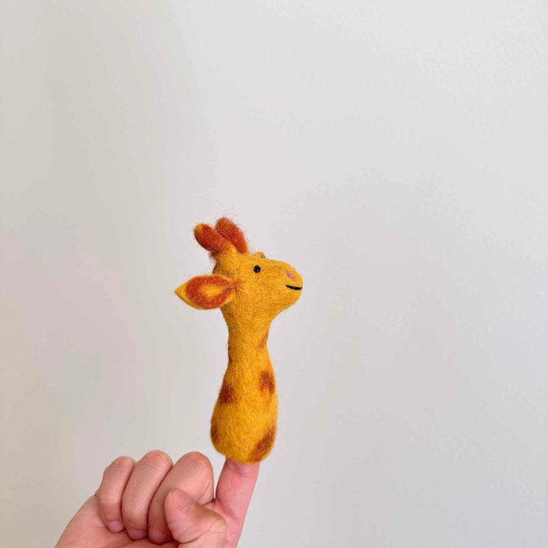 A handcrafted needle-felted giraffe finger puppet is displayed on a female hand's index finger in a close-up shot. This unique felt toy features a yellow body with distinct brown spots, small ears, and horns, showcasing its detailed fair-trade craftsmanship.