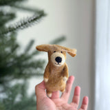 A golden-brown wool felt golden retriever finger puppet is held by a female hand against a blurred green Christmas tree and a neutral wall. This handmade felted finger puppet features small ears, a black nose, and white markings on its paws and face. Its soft, needle-felted texture creates a plush and expressive appearance.