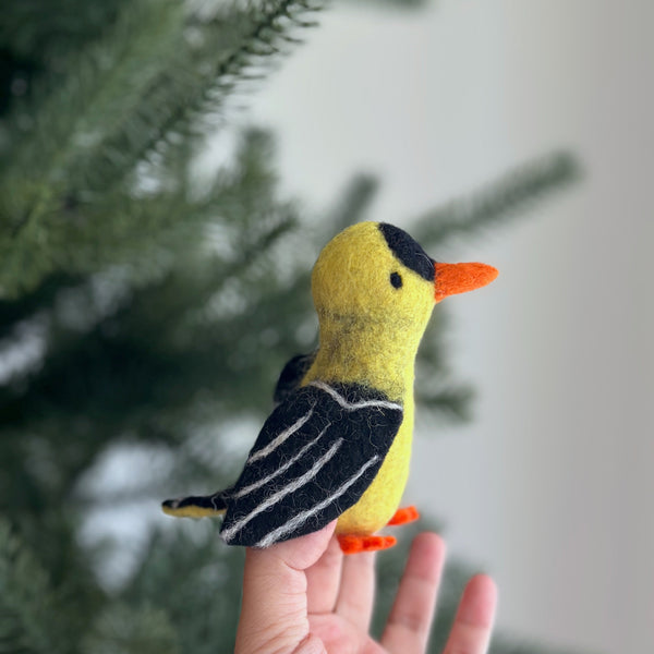 A handcrafted needle-felted goldfinch finger puppet is held gently by a female hand, with a blurred hint of a Christmas tree in the background. This charming wool felt finger puppet is predominantly vivid yellow with distinct black markings on its wings and head, complemented by a small orange beak and feet. Its detailed craftsmanship highlights the soft textures of the ethically made felt, perfect for imaginative play.